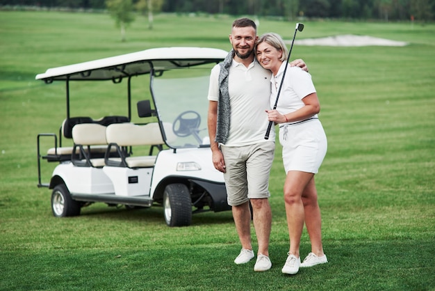 Couple of two golfers staying on the green lawn with cart behind of them.