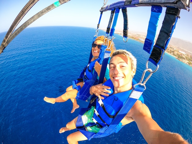 Couple of two happy people enjoying summer and vacations doing extreme activity on the sea with a boat - beautiful people taking a selfie while doing parascending together