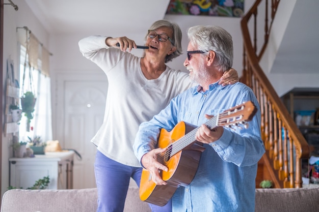 Couple of two happy seniors or mature and old people singing and dancing together at home indoor. Retired man playing the guitar while his wife is singing with a remote control of TV.