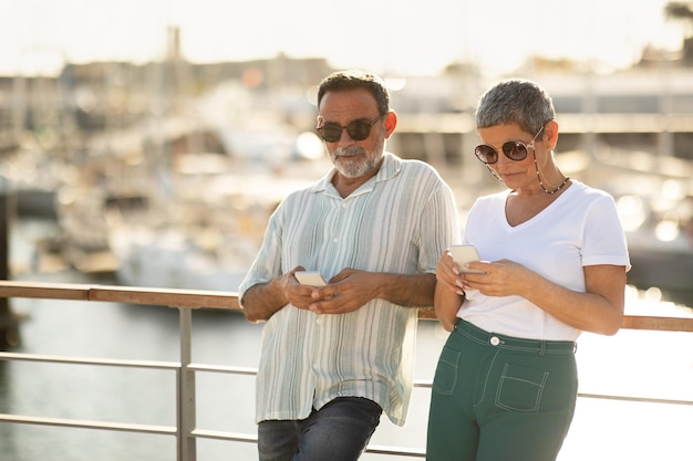 Couple using smartphones planning holiday with travel applications at marina