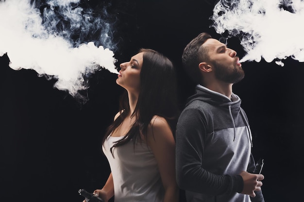 Couple vaping. Young man and woman blowing smoke at black studio background. Relationship and vape addiction concept with copy space