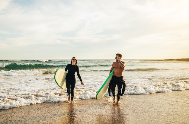 Photo a couple walks along a sandy beach after surfing both carrying surfboards they are wearing wetsuits