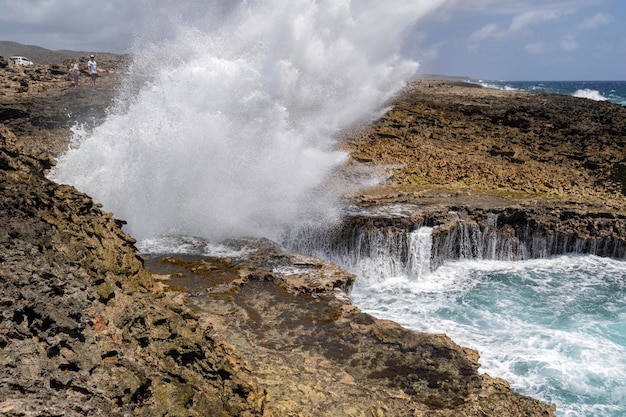 A couple watching a wave crash against a cliff with ocean spray coming off the rocks in Boka Pistol