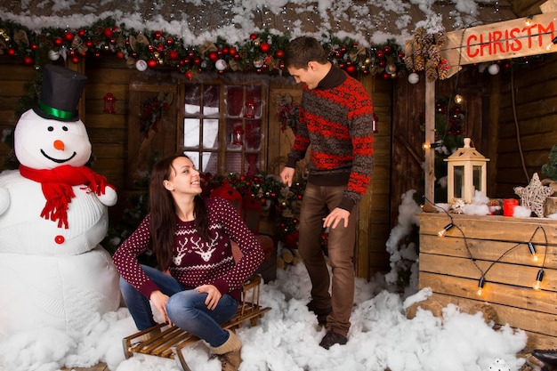 Couple Wearing Sweaters with Sled Outdoors in Winter in front of Log Cabin