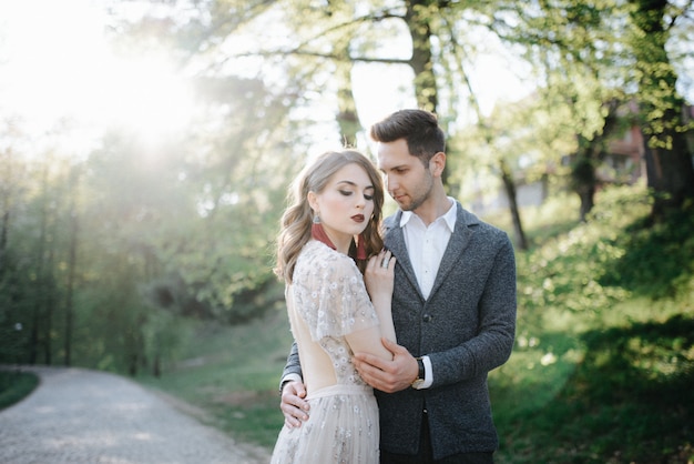 Couple in wedding attire with a bouquet of flowers and greenery is in the hands against the backdrop of the field at sunset, the bride and groom