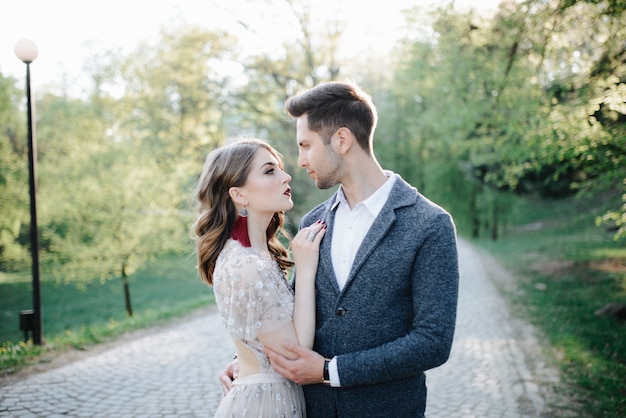 Couple in wedding attire with a bouquet of flowers and greenery is in the hands against the backdrop of the field at sunset, the bride and groom