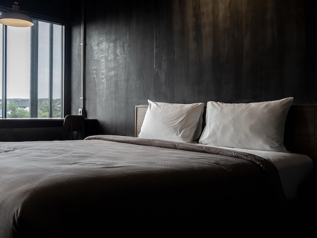 Couple white pillow on the empty bed with brown duvet on dark grunge concrete wall background near the window in hotel bedroom loft style Two soft and clean comfort pillows preparing for the guest