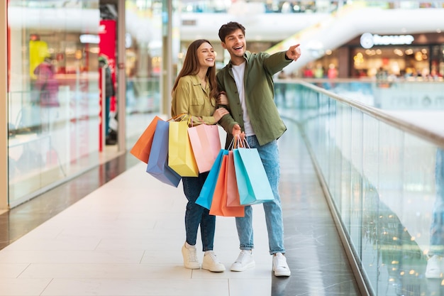 Couple with colorful bags shopping pointing finger aside in mall