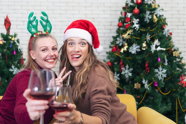 Couple with glasses of red wine near fireplace. Beautiful caucasian young woman in sweater and with santa hat on head sitting on sofa next to christmas tree