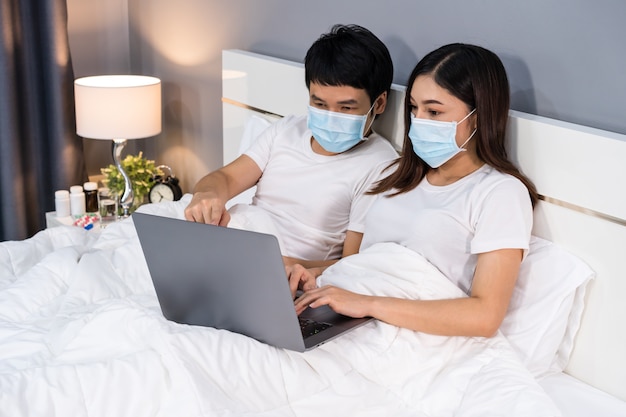 Couple with medical masks using laptop on bed during quarantine pandemic coronavirus stay at home.