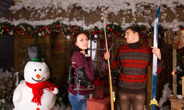 Couple with Skates and Skis Standing next to Snowman in front of Log Cabin