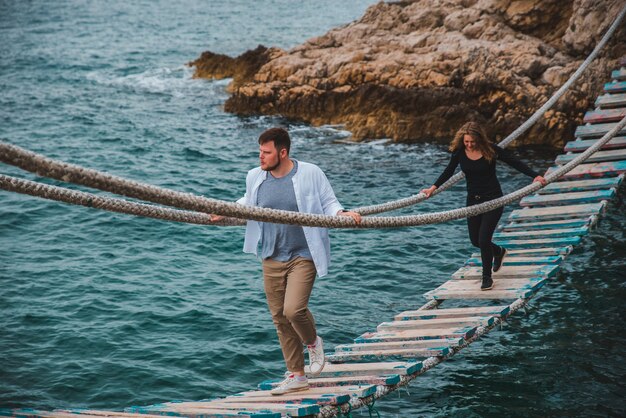 Couple woman with man walking by shaky suspension bridge crossing sea bay having fun