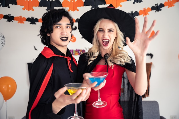 Couple young caucasian man and woman in vampires and witch clothing holding champagne glass