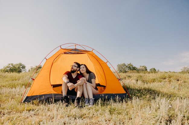 Couple of young man and woman sitting in an orange tent in mountains.