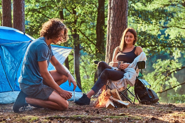 Couple of young students are chilling near bonfire in the green forest. There are tent at background.