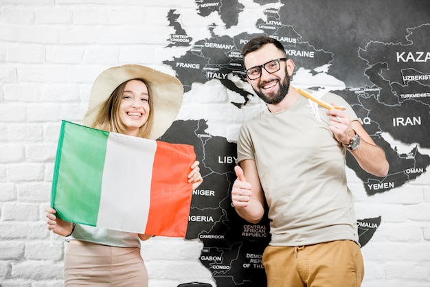 Couple of young travelers standing with italian flag near the wall with world map, dreaming about summer vacations in Italy