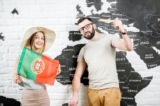 Couple of young travelers standing with portuguese flag near the wall with world map, dreaming about summer vacations in Portugal