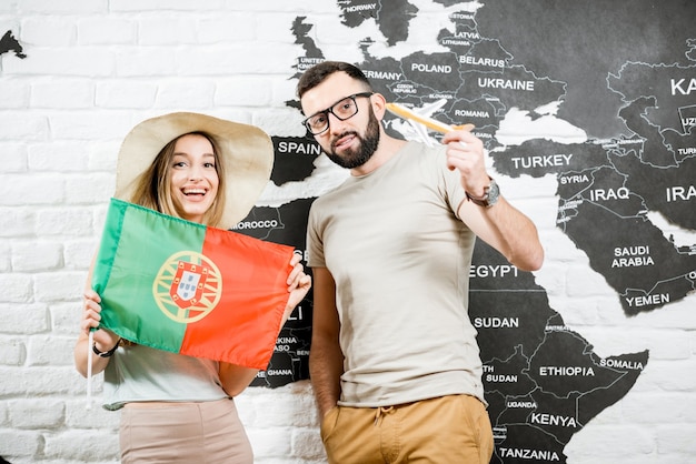 Couple of young travelers standing with portuguese flag near the wall with world map, dreaming about summer vacations in Portugal