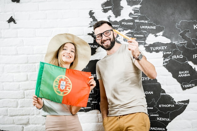 Couple of young travelers standing with portuguese flag near the wall with world map, dreaming about summer vacations in Portugal