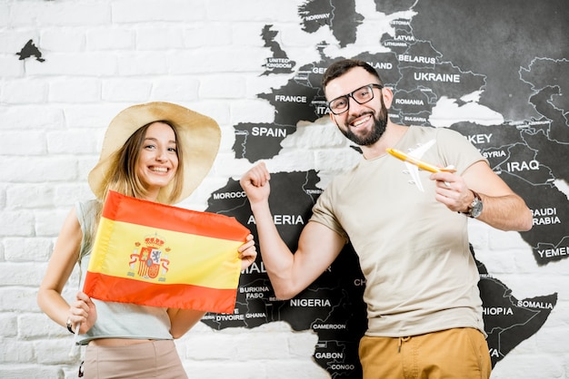 Couple of young travelers standing with spanish flag near the wall with world map, dreaming about summer vacations in Spain