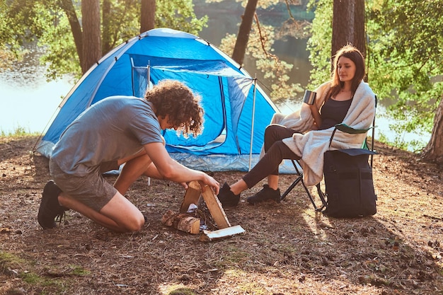 Couple of young travellers spending day in sunny forest near tent. Girl is sitting on chair, man is making bonfire.