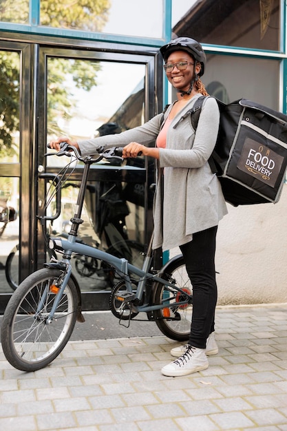 Courier in bicycle helmet waiting for customer outdoors portrait, african american woman standing near office building entrance, looking at camera. Takeaway food delivery service