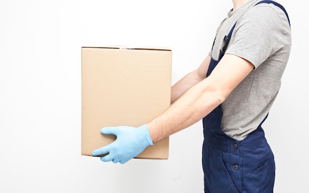 The courier is holding the carton box in his hand wearing  protective latex gloves on the gray wall