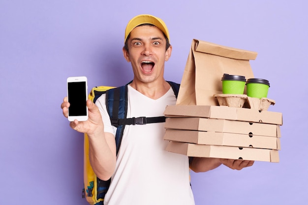 Courier man wearing white Tshirt with thermo backpack holding pizza boxes and coffee in disposable cups isolated over purple background showing cell phone with empty display for advertisement