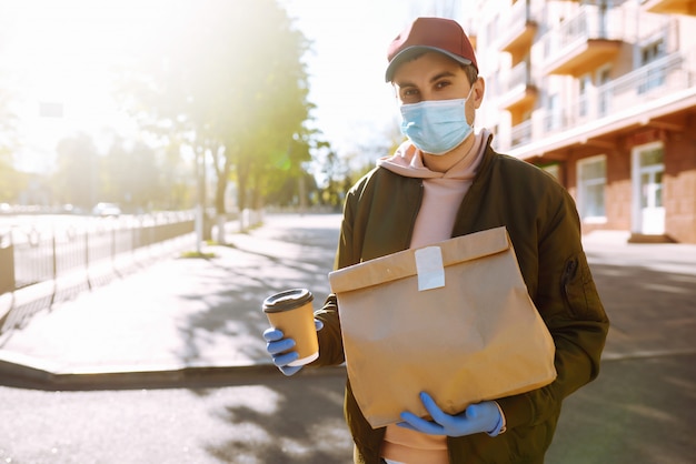 Courier in protective mask and medical gloves on a scooter delivers craft paper packet with food. Delivery service under quarantine, disease outbreak, coronavirus covid-19 pandemic conditions.