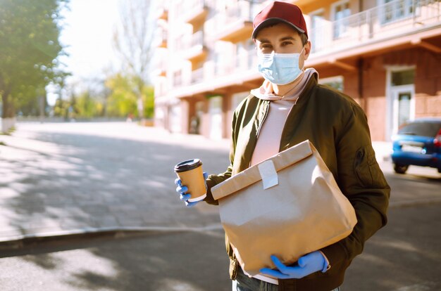 Courier in protective mask and medical gloves on a scooter delivers craft paper packet with food. Delivery service under quarantine, disease outbreak, coronavirus covid-19 pandemic conditions.