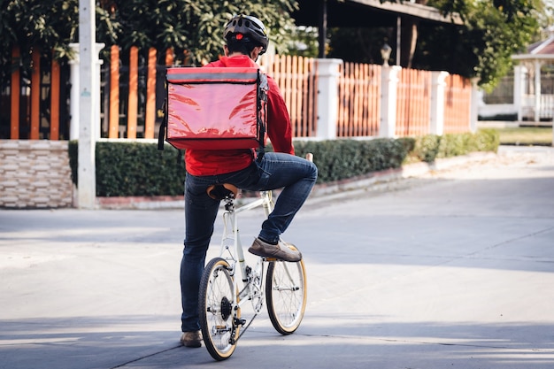 Courier in red uniform with a delivery box on back riding a bicycle and looking on the cellphone to check the address to deliver food to the customer. Courier on a bicycle delivering food in the city.