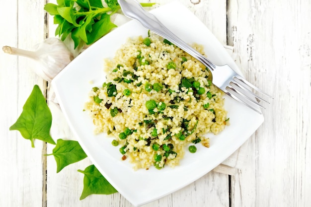 Couscous with spinach and green peas in a plate on a kitchen towel, basil and fork on a wooden plank background