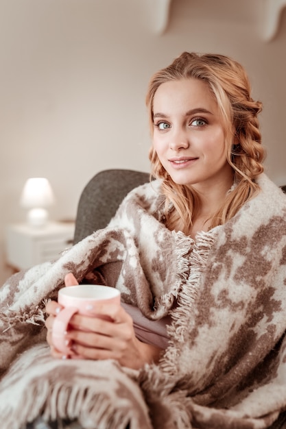 Covered in blanket. Alluring long-haired woman with big blue eyes and flawless glowing skin sitting in cozy armchair