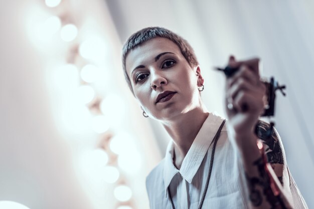 Photo covered in tattoos. unusual young lady with boyish haircut keeping ink machine and looking monumental and scary