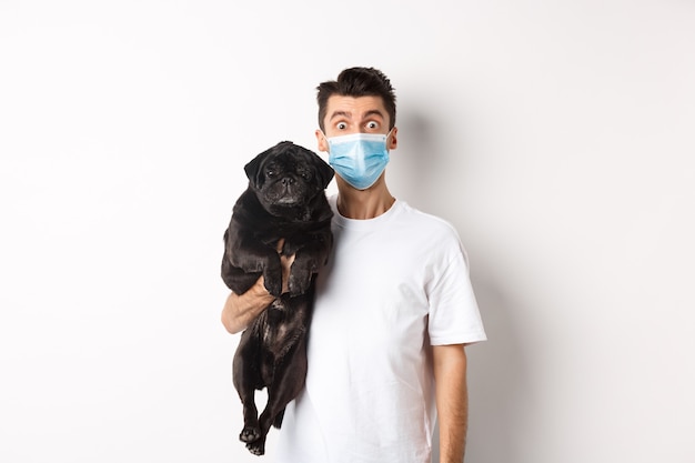 Covid-19, animals and quarantine concept. Funny young man in medical mask holding cute black pug dog, standing over white background