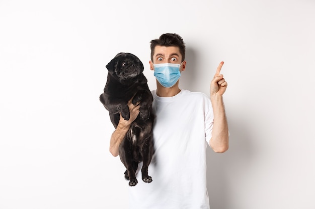 Covid-19, animals and quarantine concept. Young man in medical mask holding cute black pug, dog looking right and owner pointing at logo, white