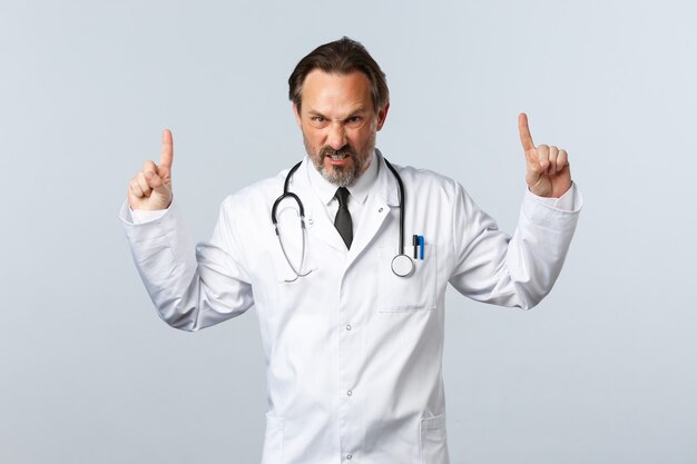 Covid-19, coronavirus outbreak, healthcare workers and pandemic concept. Angry outraged male doctor in white coat, pointing fingers up grimacing furious, looking mad at camera, white background