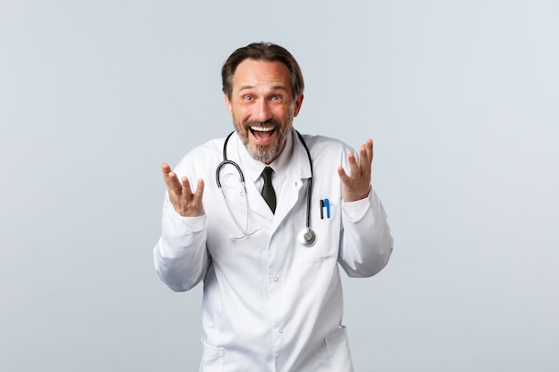 Covid-19, coronavirus outbreak, healthcare workers and pandemic concept. Excited happy male doctor laughing and explaining something funny, smiling broadly, standing white background