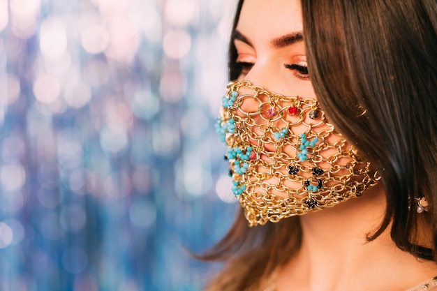 Covid-19 fashion. Pandemic Xmas. Quarantine party accessory. Profile portrait of sensual woman in ethnic style gold chain face mask on blue pink bokeh light blur tinsel copy space background.