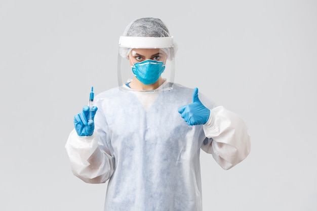 Covid-19, medical research, healthcare workers and quarantine concept. Confident professional doctor in personal protective equipment PPE costume, show thumb-up, hold syringe with corona vaccine