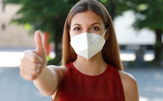 COVID-19 Optimistic girl wearing protective mask  FFP2 avoiding Coronavirus disease 2019 showing thumbs up in city street