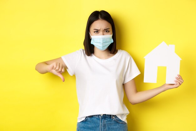 Covid-19 and real estate concept. Disappointed asian woman in medical mask, showing thumb down and paper house cutout, standing upset against yellow background.