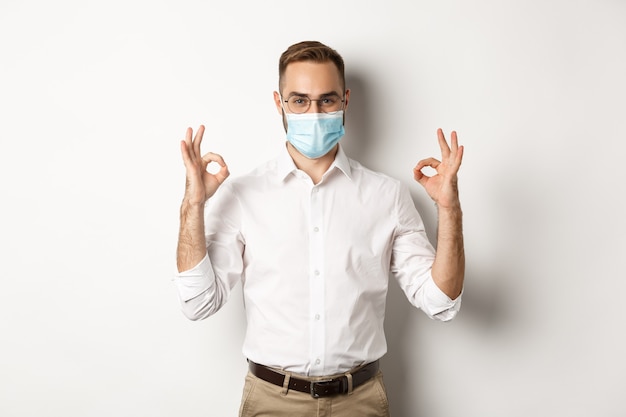 Covid-19, social distancing and quarantine concept. Confident businessman wearing medical mask and showing okay signs in approval, white background.