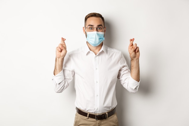 Covid-19, social distancing and quarantine concept. Hopeful employee in medical mask, holding fingers crossed and making wish, white background.