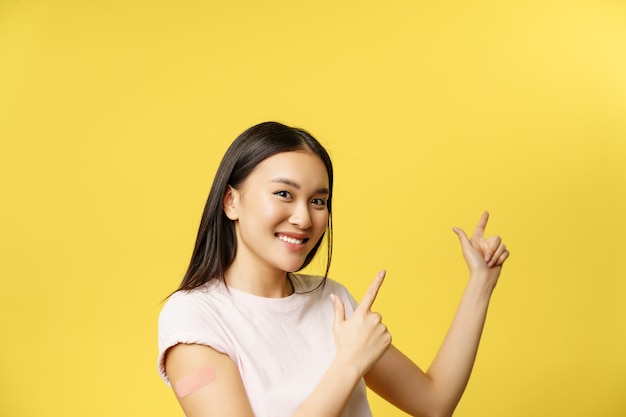 Covid-19 vaccination concept. Cute vaccinated asian woman demonstrating banner, pointing fingers at empty space for promotional text, yellow background