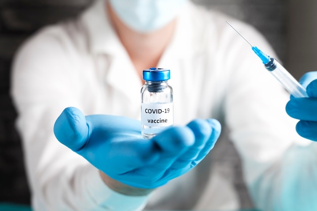 COVID-19 vaccine in researcher hands, male doctor in a white coat, in a medical mask, in blue gloves holds syringe and bottle with vaccine for coronavirus cure. Concept of corona virus treatment.