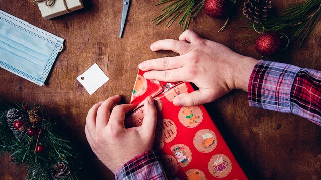 Covid closeup shot of hands of a man packaging a Christmas present with a medical mask next to him