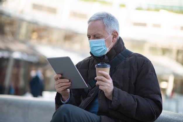 Covid coronavirus lifestyle masked elder man using his tablet while sitting outdoor in a city square