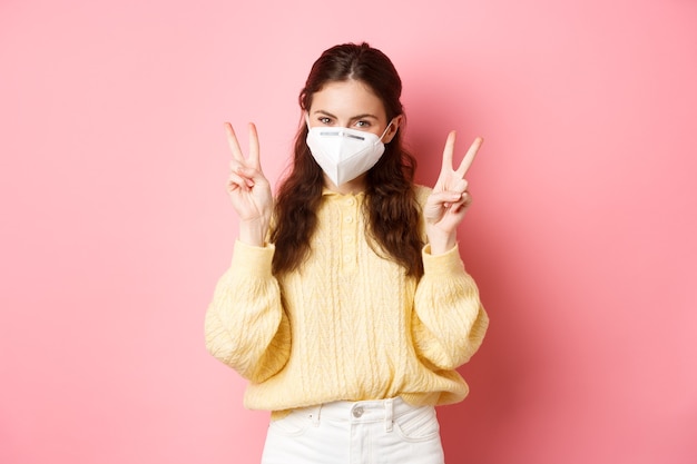 Covid lockdown and pandemic concept cute glamour girl wears medical respirator for going outdoors during quarantine shows peace vsign stands against pink wall