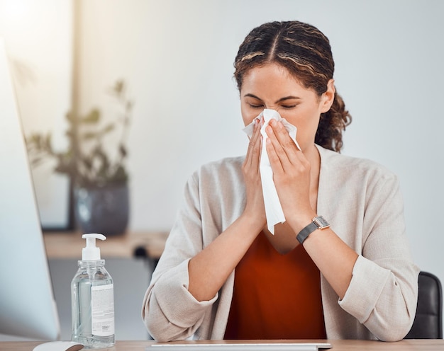 Covid sick and woman blowing her nose with a tissue while working in her modern office during pandemic Flu cold or sinus allergy sneeze of girl from mexico sitting at her desk with health problems